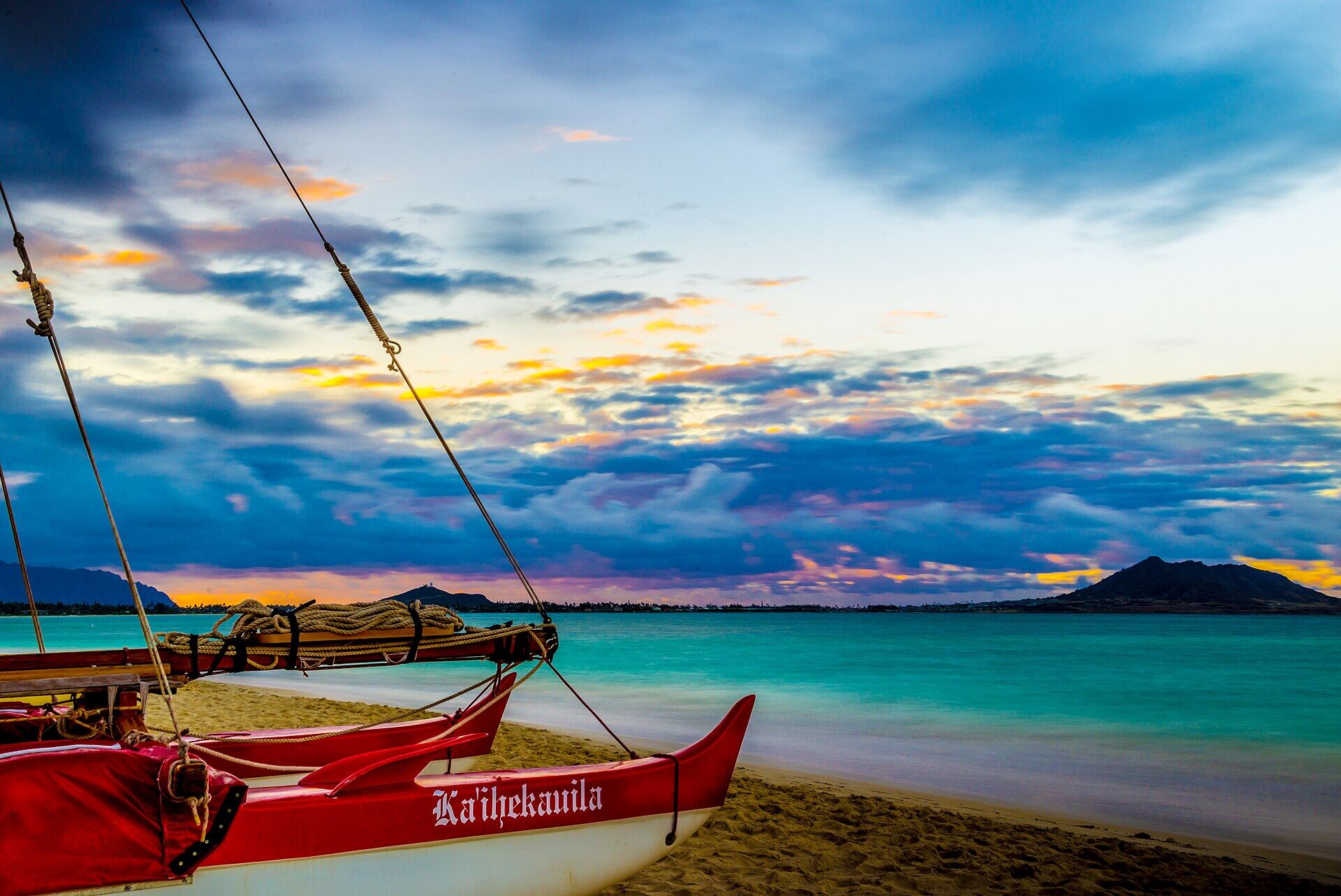 A Hawaiian sailing canoe pulled up on Kailua Beach at dusk, with the far side of the bay behind it